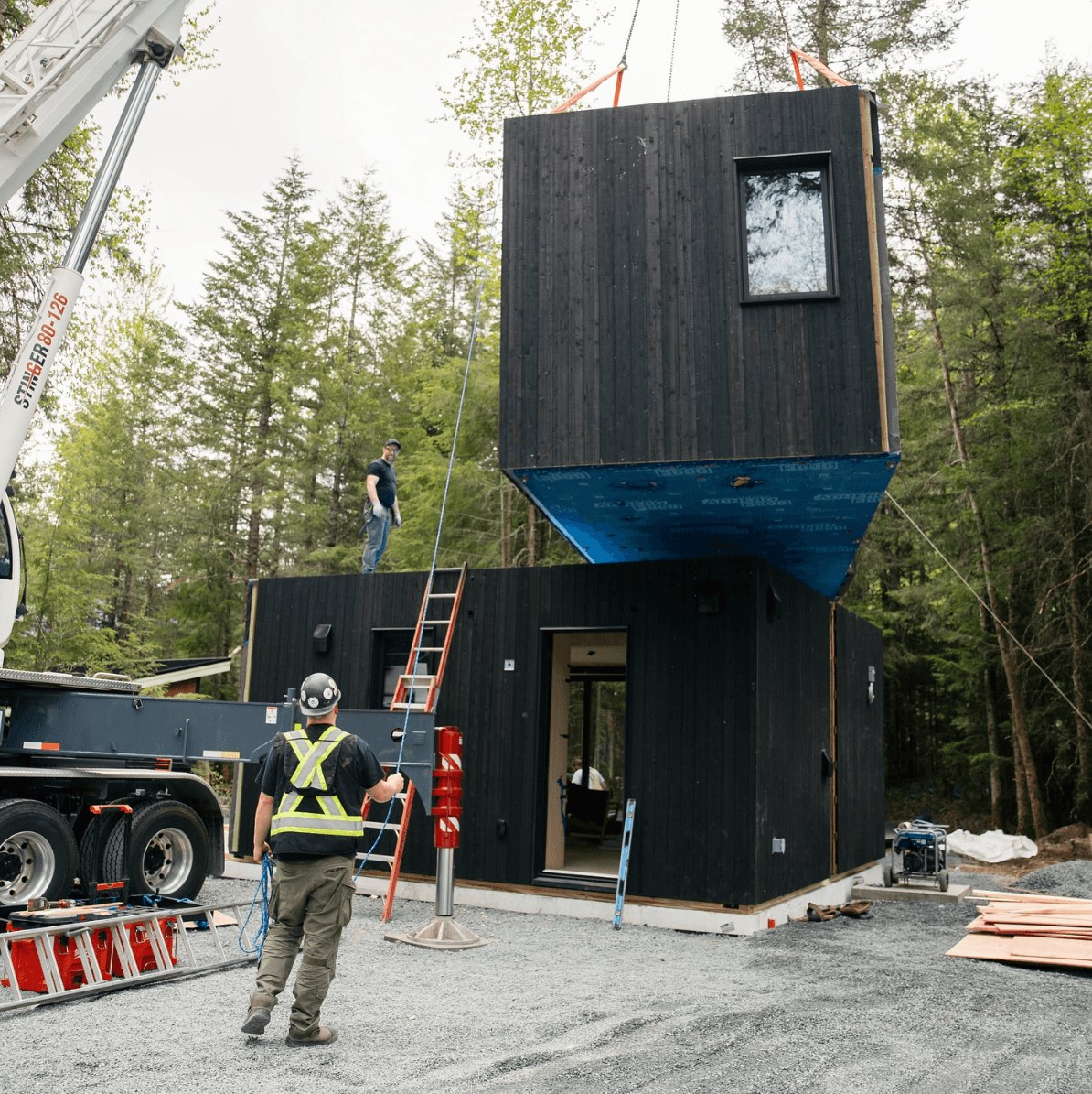 Crane placing a home module above another section at a wooded site